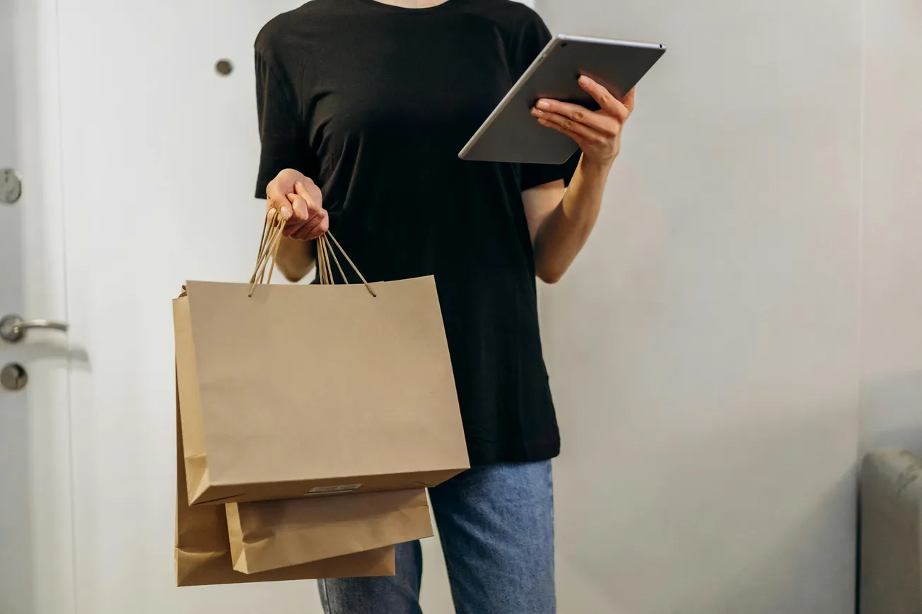 A person in a black shirt and jeans holds shopping bags and a tablet, standing indoors. The scene suggests multitasking and consumer activity.
