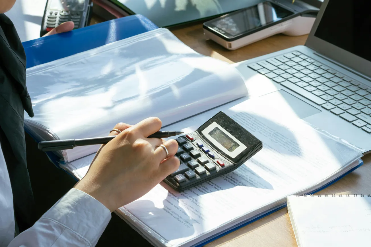 Person using a calculator and pen over open documents on a desk, near a laptop and smartphone. The scene conveys focus and productivity.