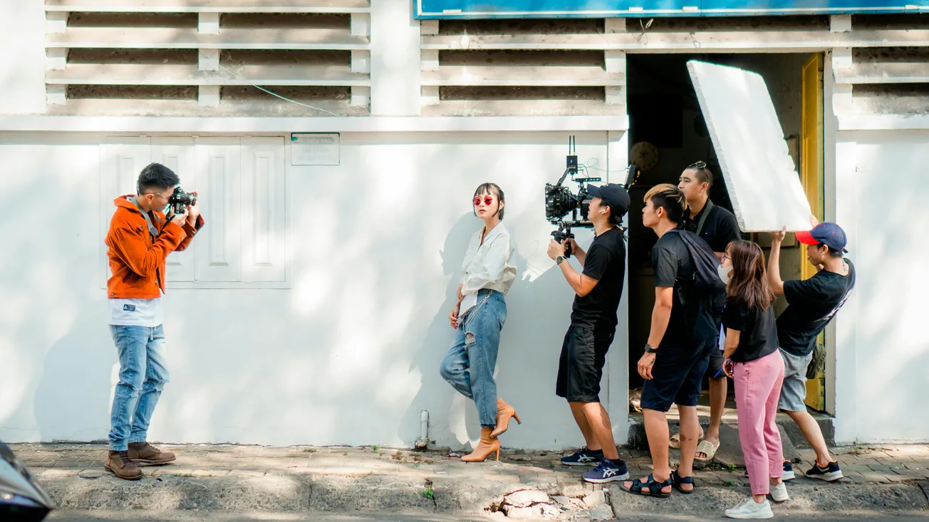 A stylistic photoshoot scene outdoors shows a photographer capturing an image of a woman against a wall, flanked by a film crew with equipment.