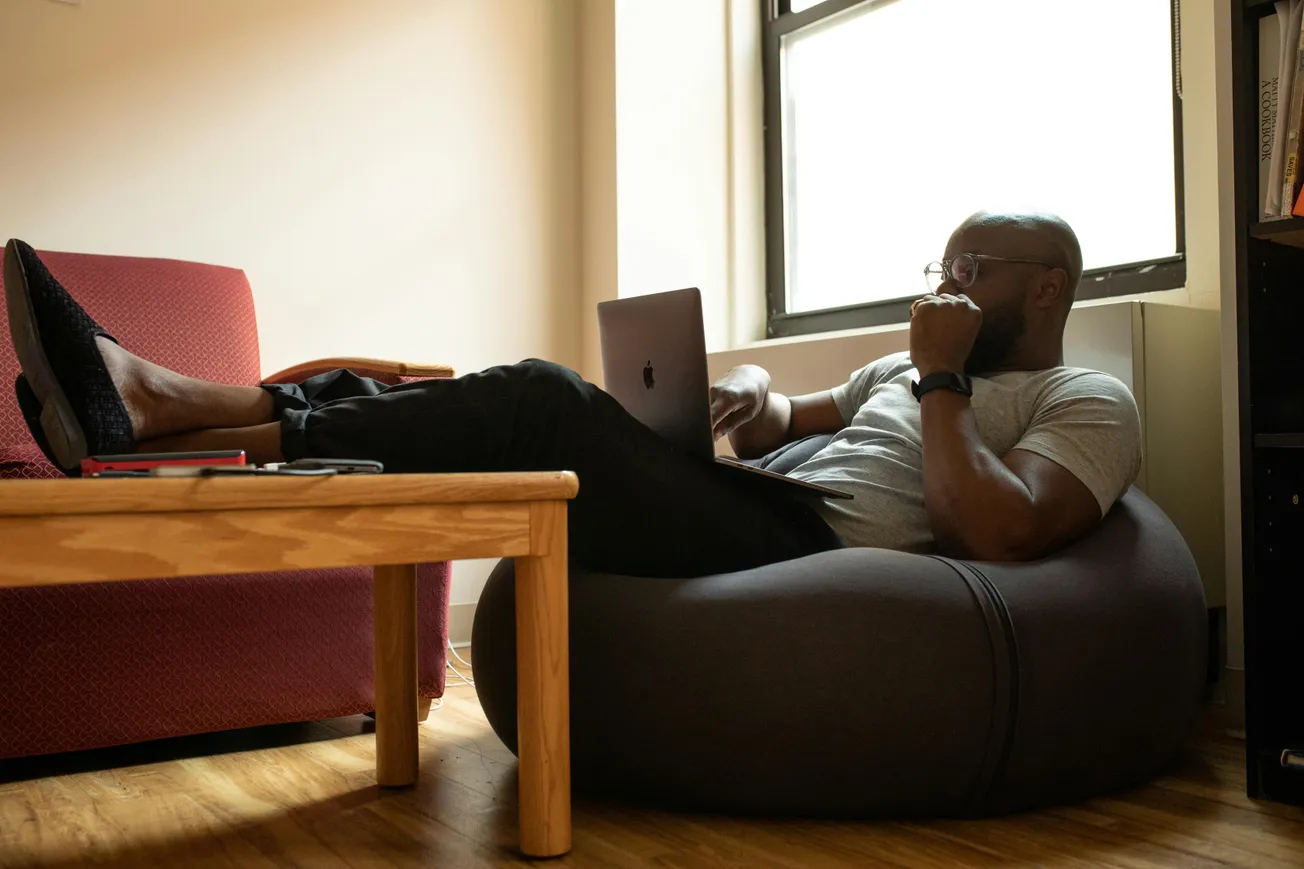 Man relaxing on a bean bag, using a laptop with feet up on a table. A casual setting with soft window light, conveying a relaxed, focused mood.