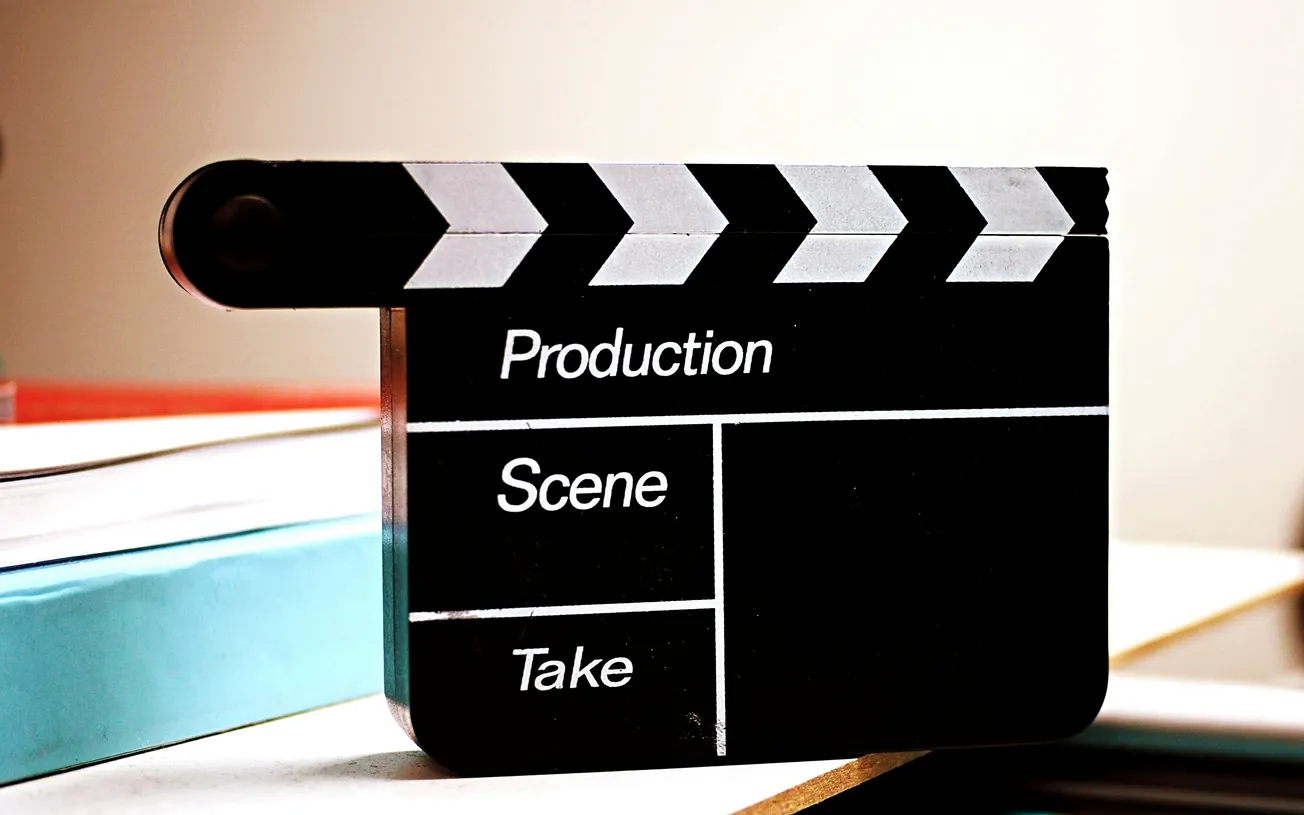 A black and white film clapperboard with the words "Production," "Scene," and "Take" sits on a table, evoking a sense of filmmaking and creativity.