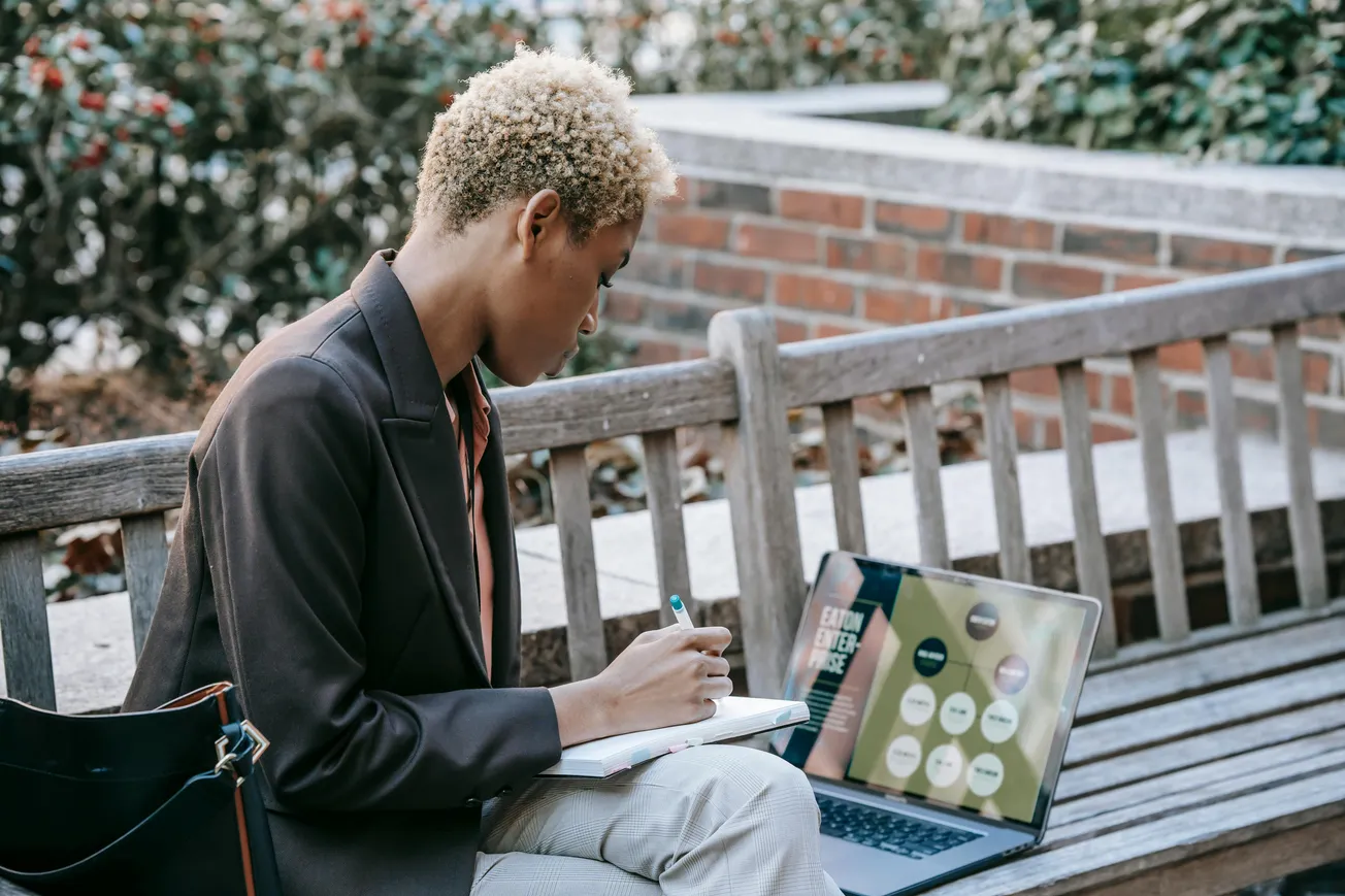 A person with short blond hair sits on a wooden bench outdoors, writing in a notebook. An open laptop beside them displays colorful charts.