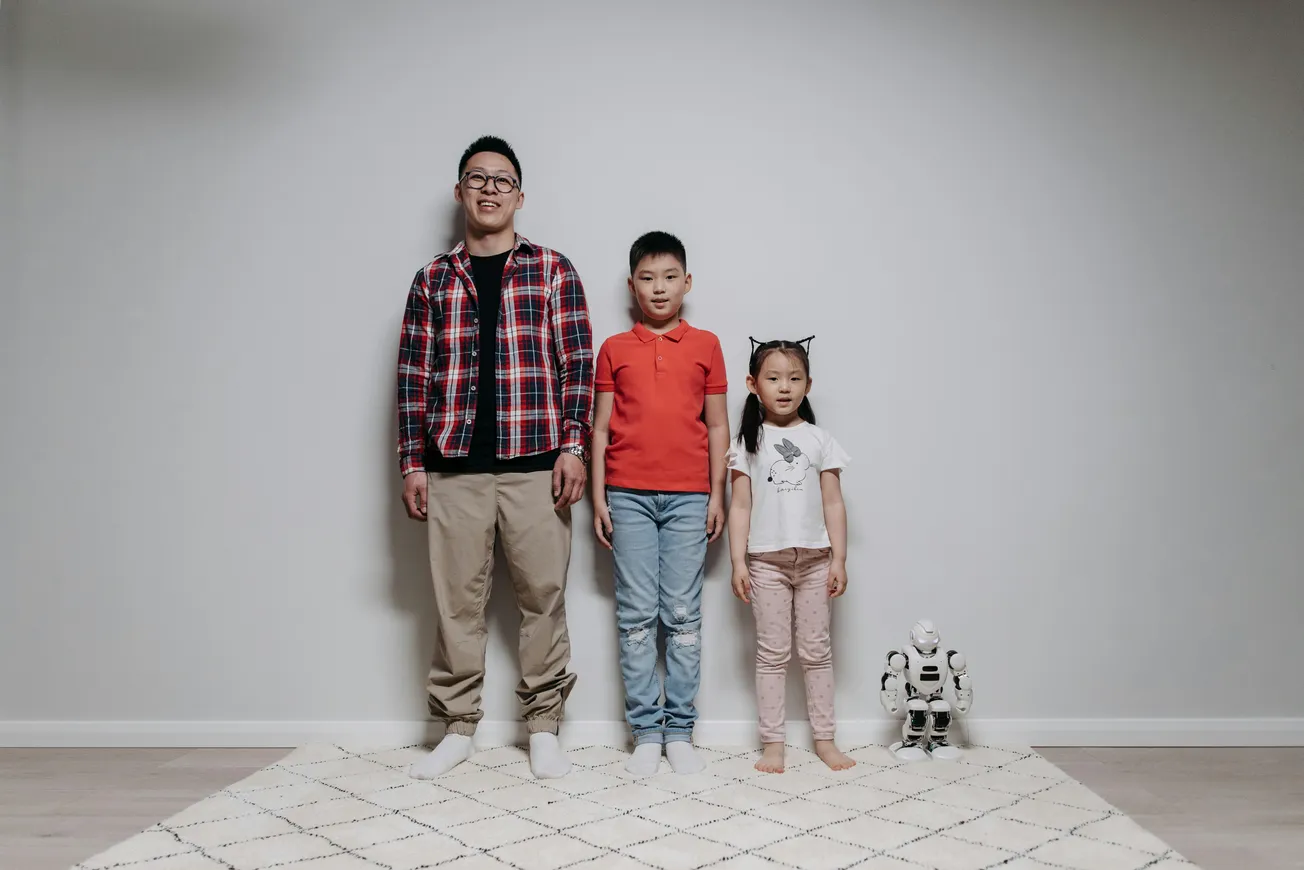 A smiling man and two children stand against a plain wall; the man wears glasses and a plaid shirt, the boy in orange, the girl in light pink. A small robot is nearby.