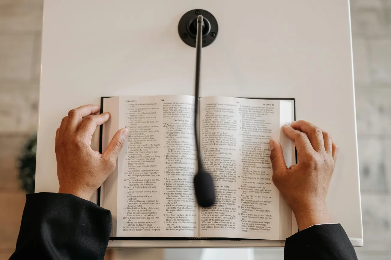 Hands hold open a Bible on a podium, with a microphone above. The scene suggests a religious setting, evoking a sense of reverence and focus.