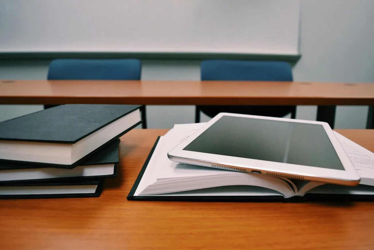 Stack of books and a tablet lie on a wooden desk in a classroom. Blue chairs and desks in the background give a studious, academic atmosphere.