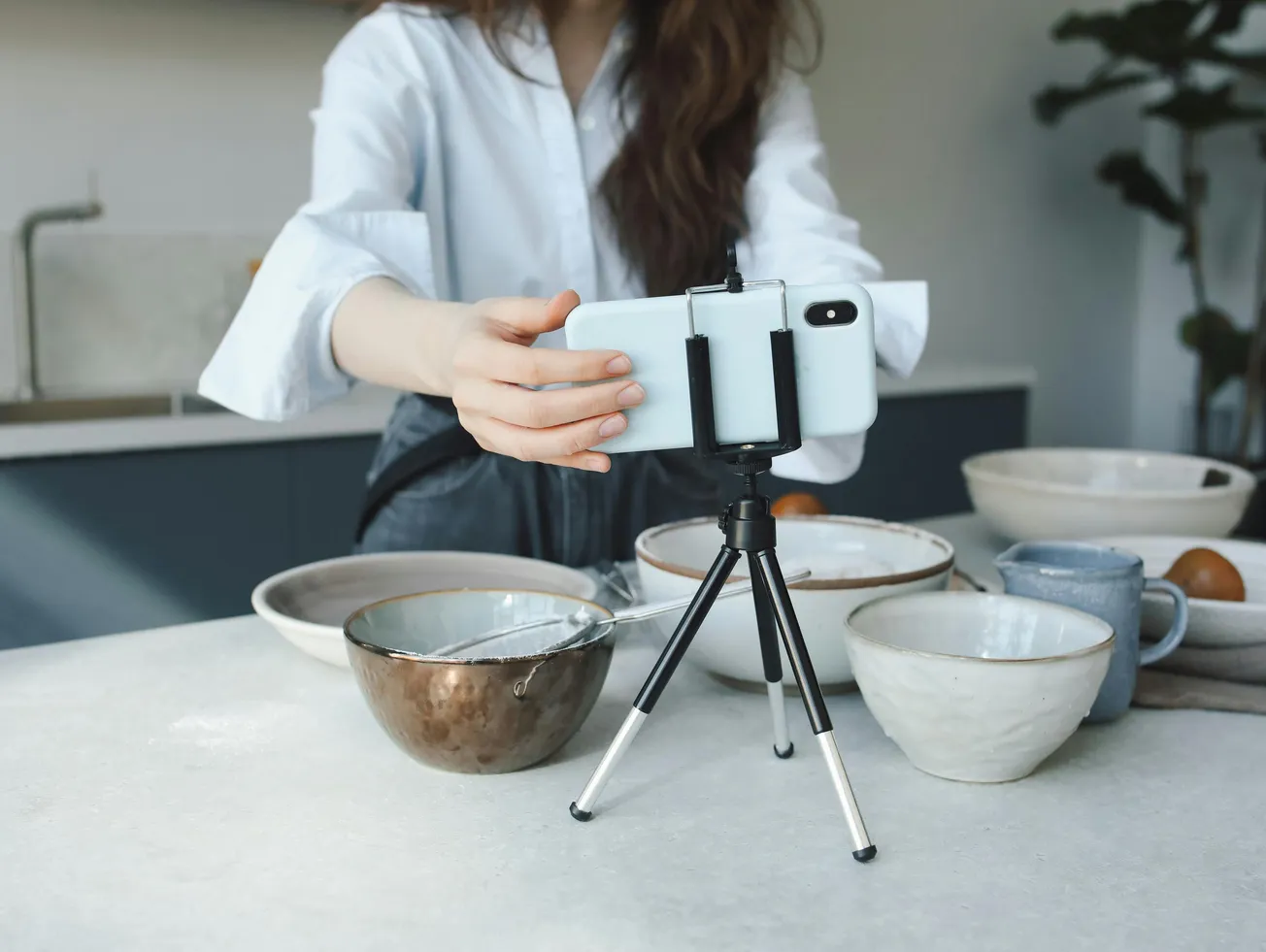 A person takes a photo using a smartphone on a tripod, capturing ceramic bowls and a mug on a kitchen counter. The scene feels calm and focused.