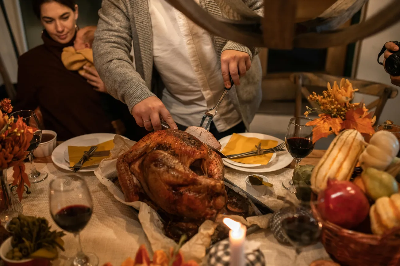 A cozy dinner scene showing a person carving a golden-brown turkey at a festive table. A woman holds a baby nearby. Autumn decor and wine glasses create a warm ambiance.