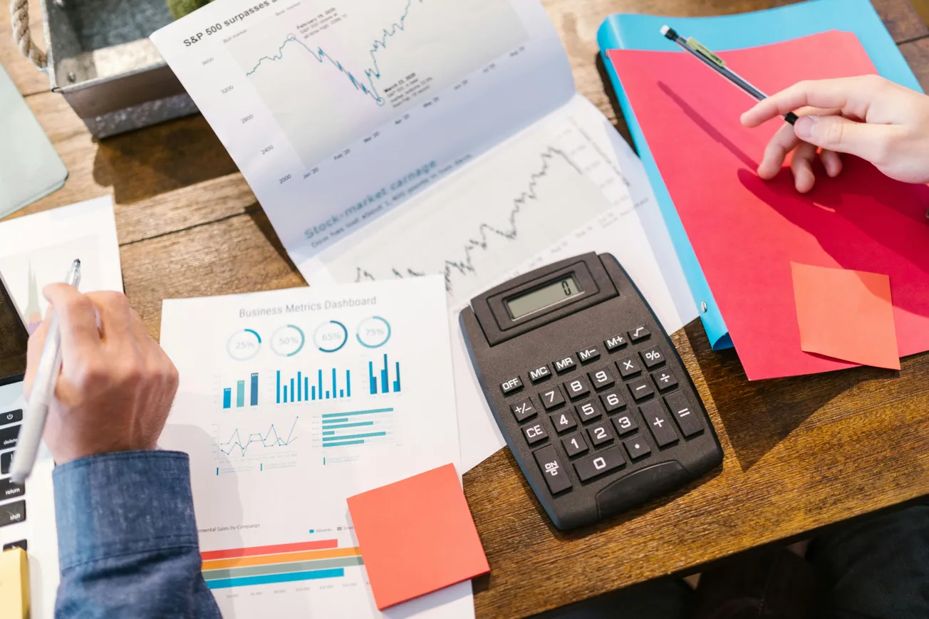 Hands working on financial documents with charts, a calculator, and colored folders on a wooden desk, conveying a busy, analytical tone.