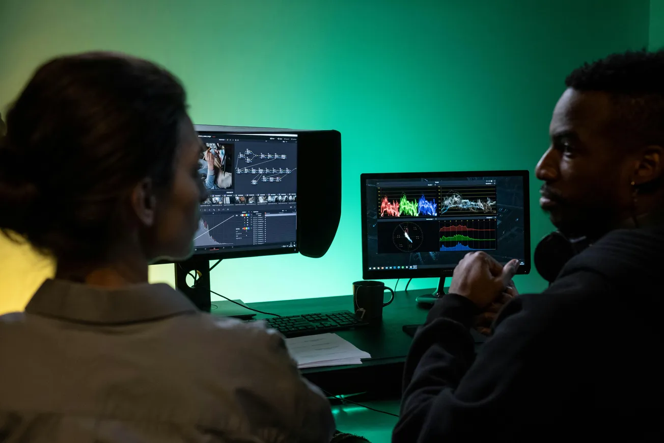 Two people sit in a dimly lit room, focused on dual monitors showing video editing software. The ambient green light sets a contemplative mood.