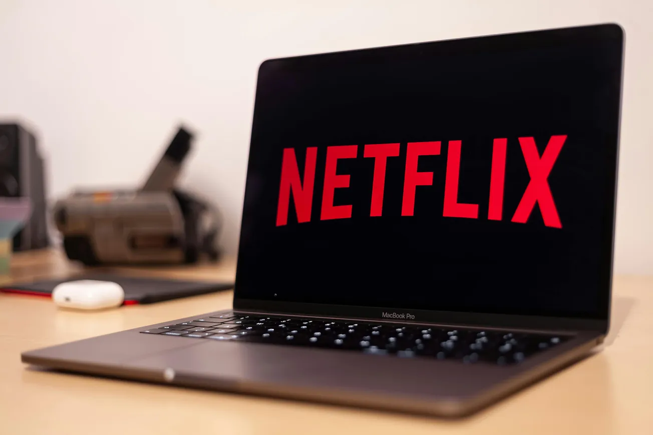 A MacBook Pro on a desk displays the Netflix logo on its screen. Nearby are an AirPods case and a blurred background with office items, suggesting a relaxed mood.