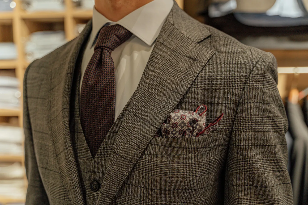 Man in a gray plaid suit with a white shirt and dark tie stands in a clothing store. A patterned pocket square adds a touch of elegance.