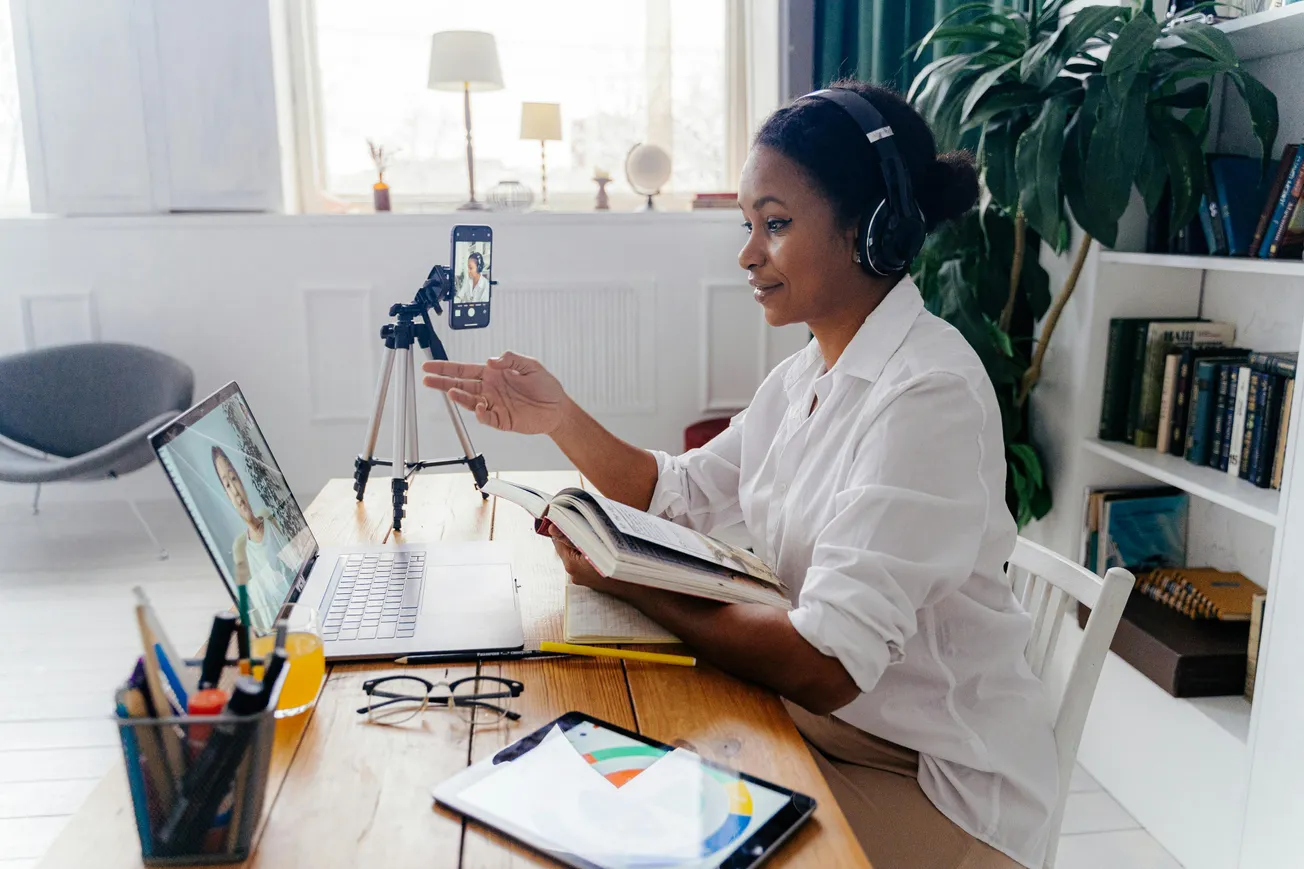 Woman in a white shirt on a video call at a home office desk. She wears headphones, holds a book, and gestures towards a laptop, conveying focus and engagement.