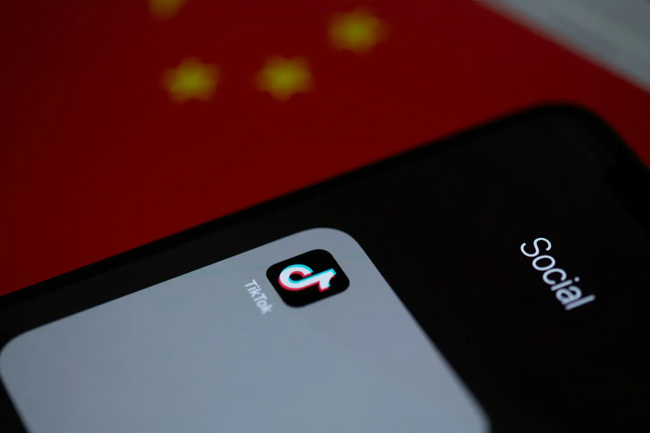 Close-up of a smartphone screen showing the TikTok app icon under a folder labeled "Social." In the background, the red flag of China is partially visible.