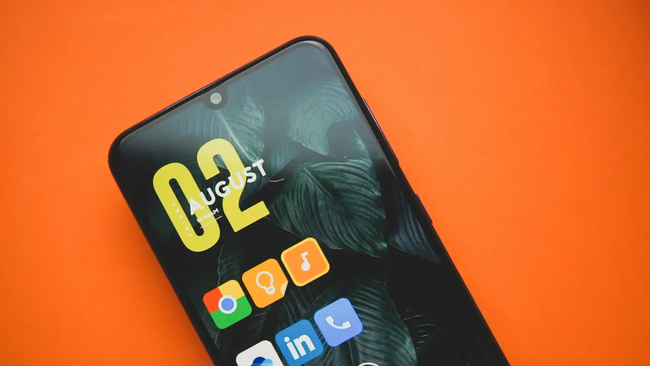 Close-up of a smartphone on an orange background displaying a date, "02 August," with app icons including Chrome and LinkedIn against a leaf-patterned wallpaper.