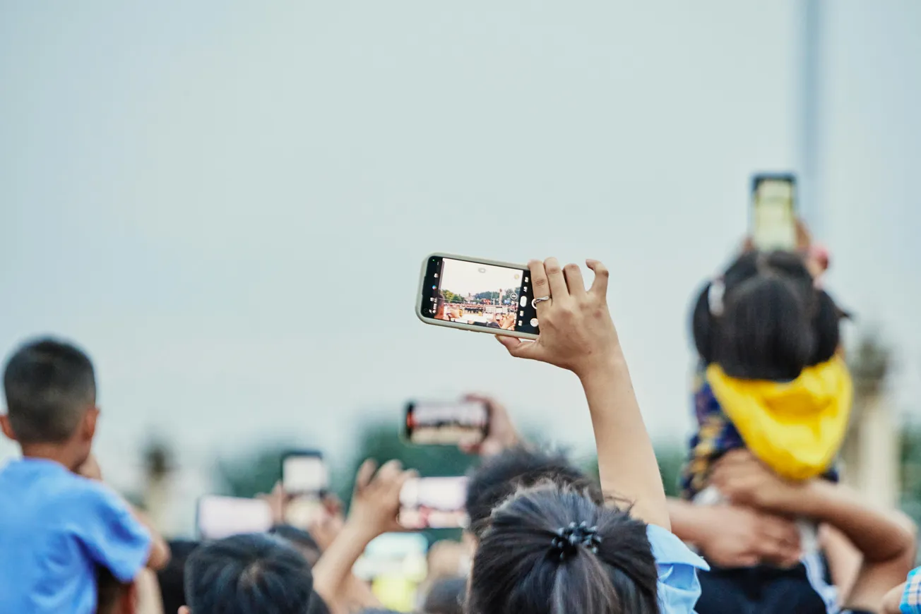 A crowd holds up smartphones, capturing a distant scene. A child on a shoulder wears a yellow hoodie. The moment feels lively and anticipatory.