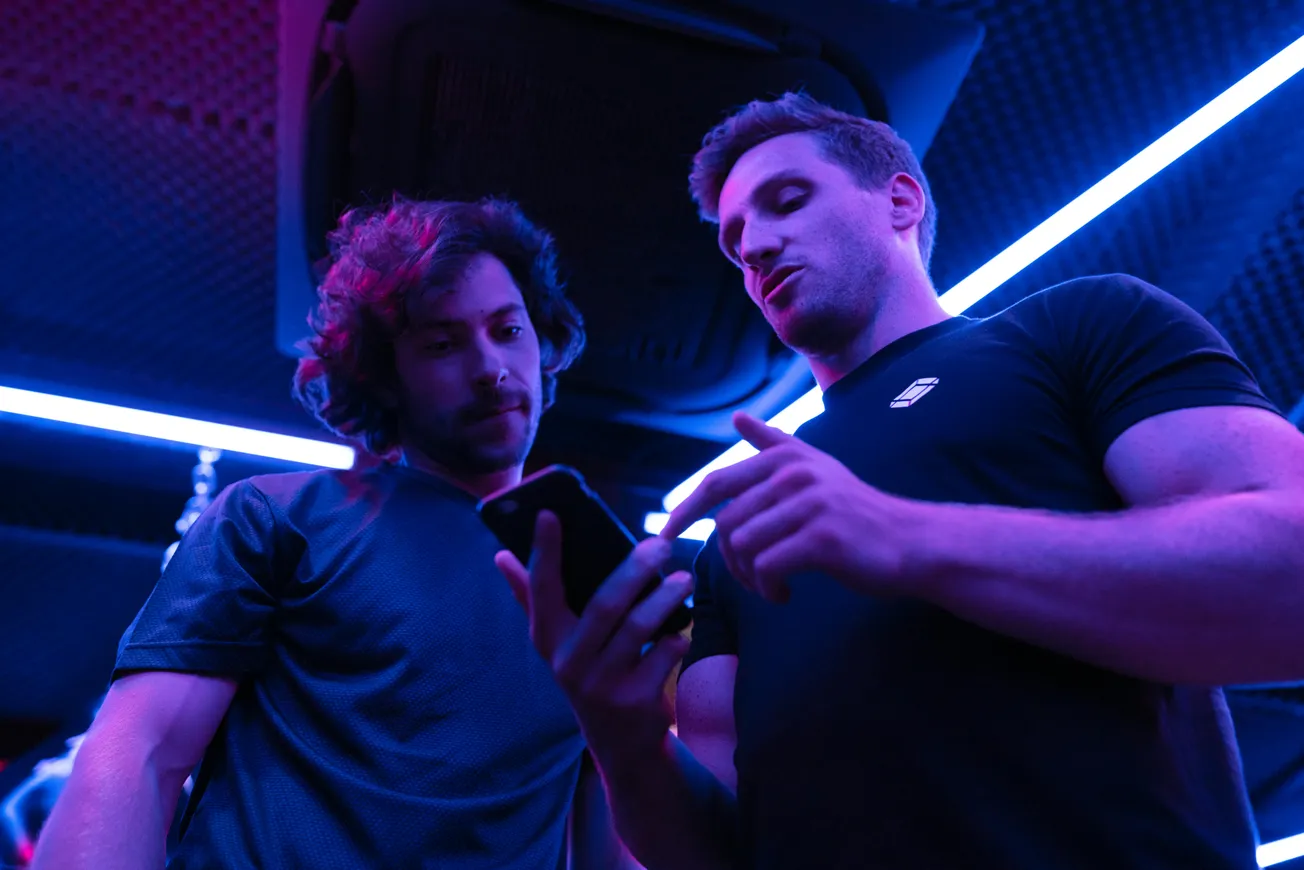 Two men in a room with vibrant purple and blue neon lighting discuss something on a smartphone, creating a modern, tech-savvy atmosphere.