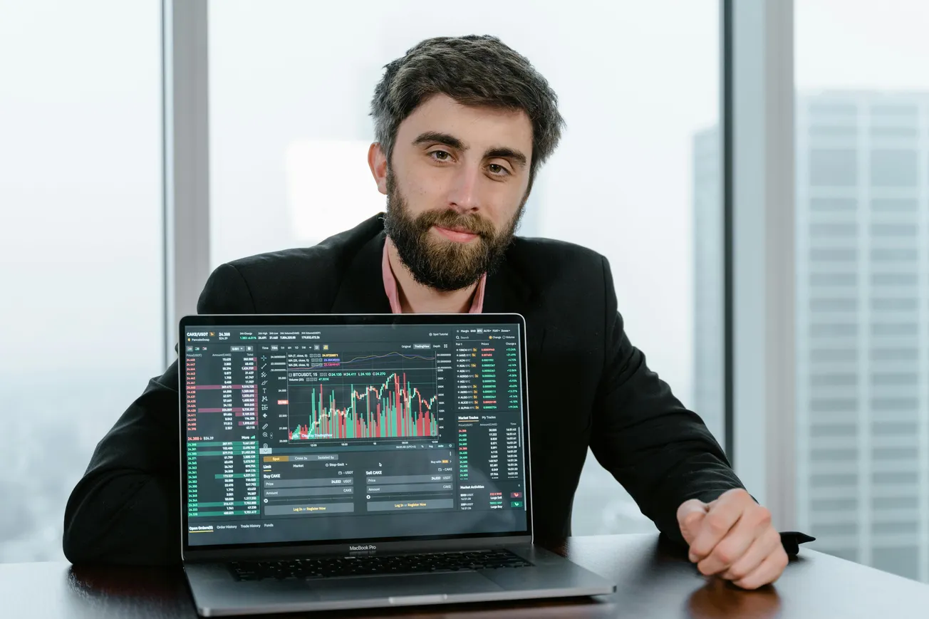 A man in a suit sits by a window, holding a laptop showing stock market graphs. The mood is professional and analytical, with a cityscape backdrop.