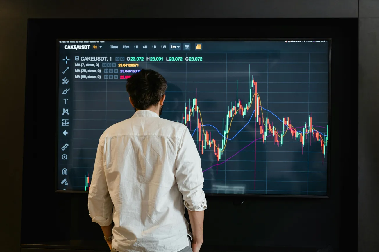 A person in a white shirt stands facing a large screen displaying a colorful financial chart with fluctuating lines, suggesting market analysis focus.