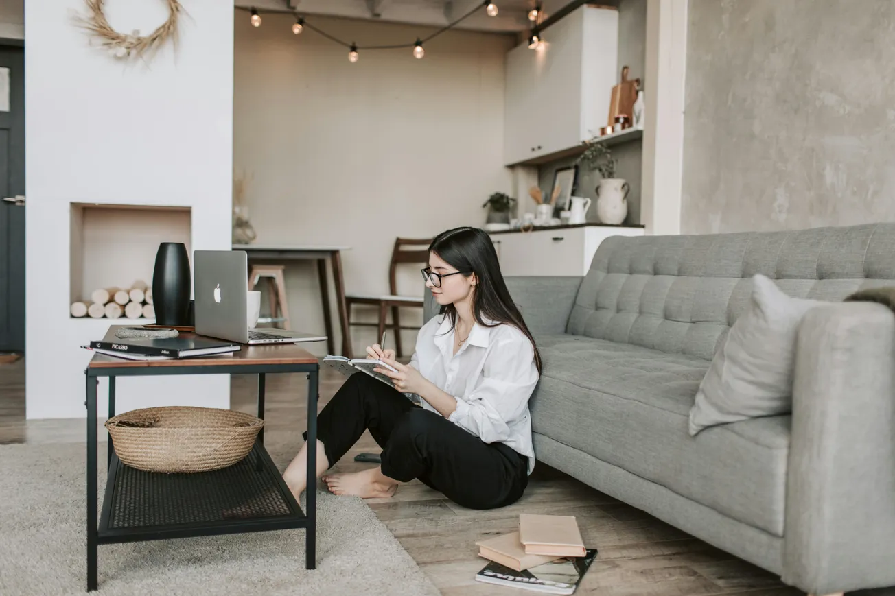 A woman sits on the floor by a gray sofa, working on a tablet. A laptop and books are on the coffee table. The room is cozy with warm lighting.