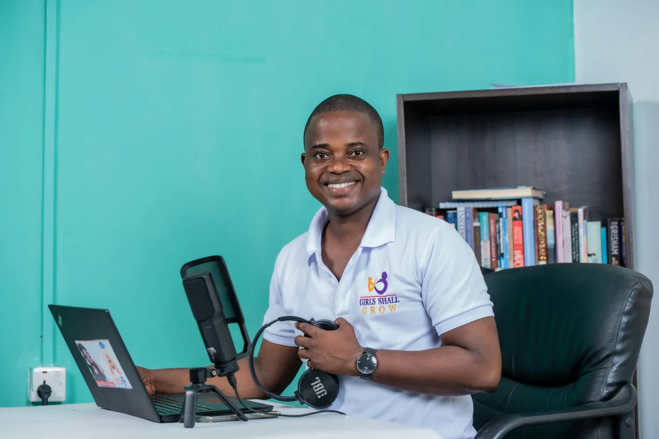 A smiling man in a white polo shirt sits at a desk with a laptop and microphone. A bookshelf with books is in the background, creating a cheerful, professional atmosphere.