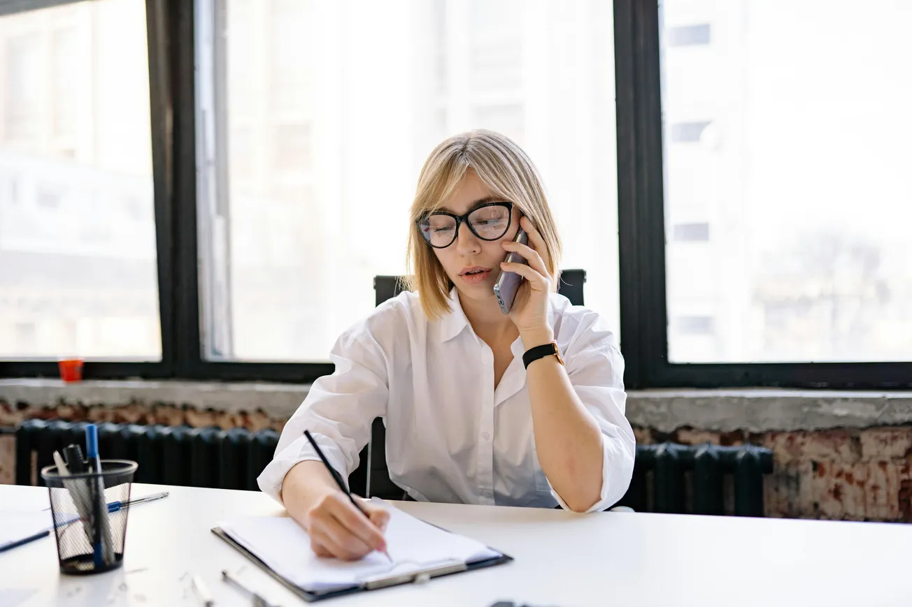 A woman in a white shirt and glasses talks on a phone, writing in a notebook at a desk. Bright window background conveys a focused work mood.