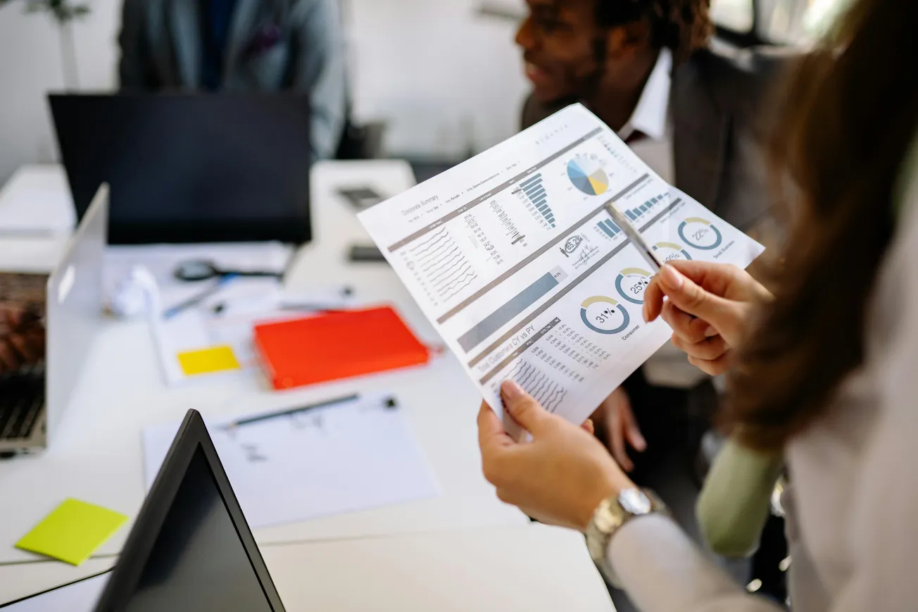 Two people in a meeting analyze financial charts on a paper. Laptops, notes, and a bright workspace suggest a collaborative, focused atmosphere.