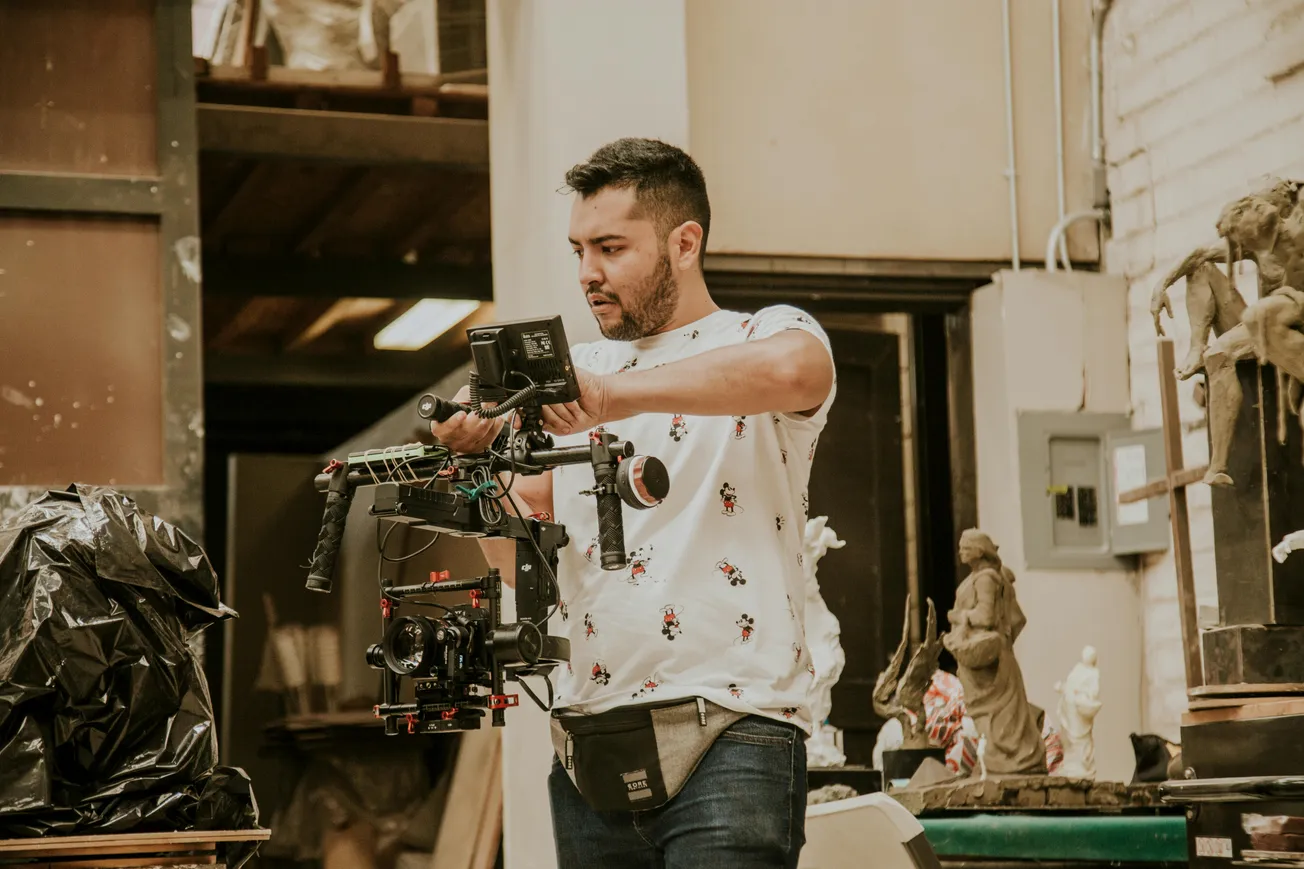 Man in a white patterned shirt focuses intently while adjusting a camera rig in a cluttered workshop, surrounded by sculptures and artistic tools.
