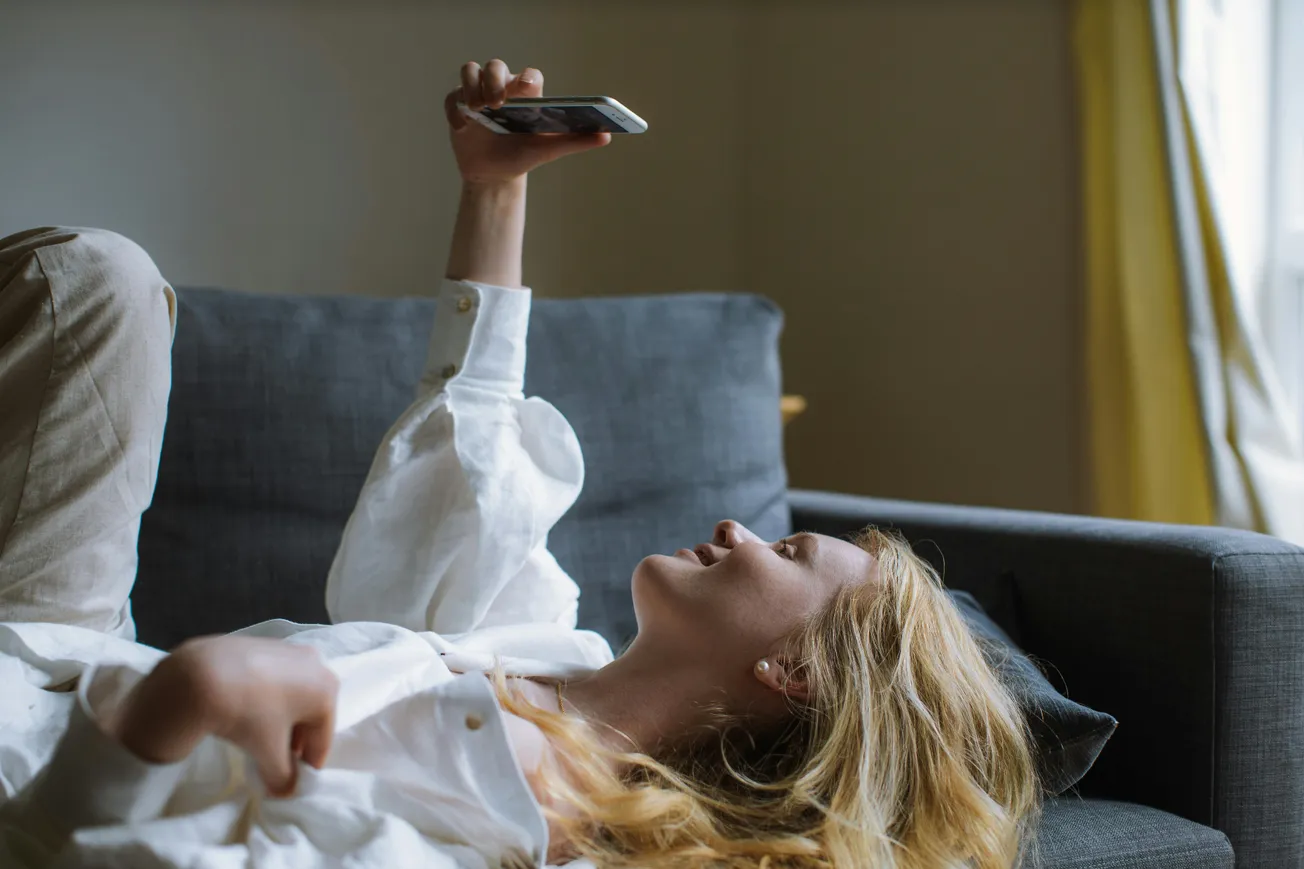 A woman lounging on a gray sofa holds a phone above her. She wears a white shirt, appearing relaxed and content. Natural light filters through a window.