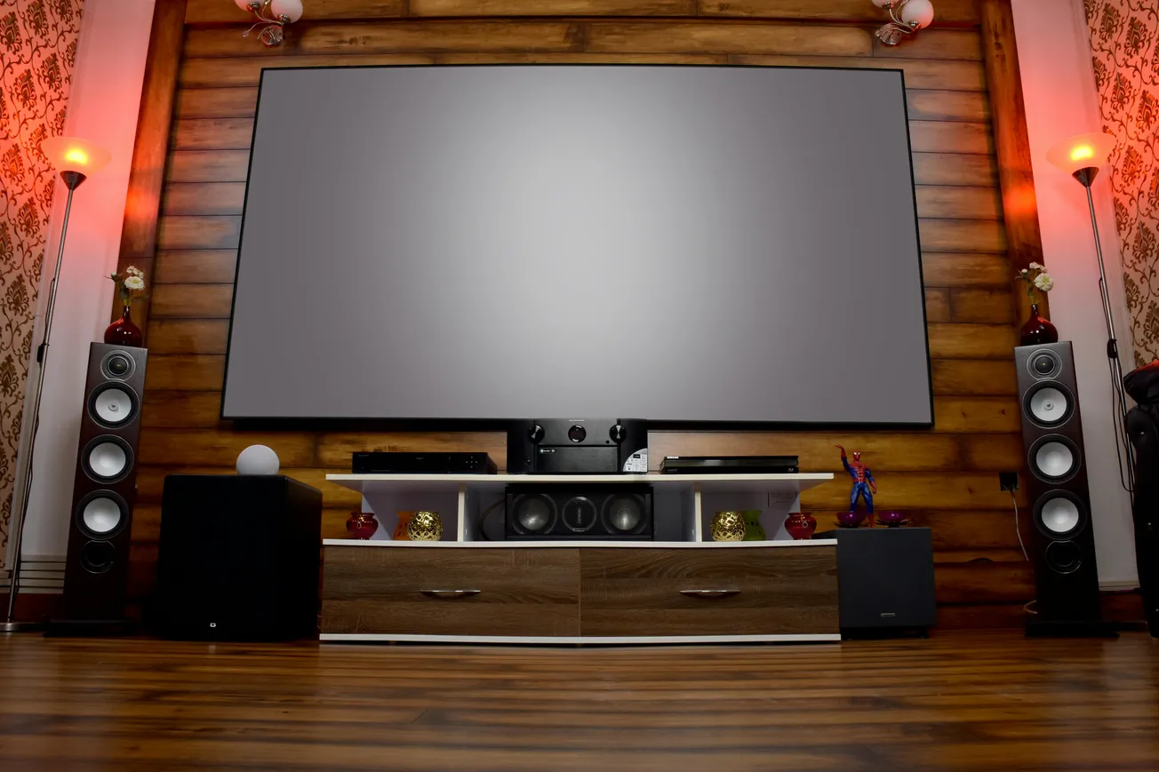 Home theater room with a large screen, wooden wall, and floor. Two tall speakers and red-lit lamps flank the setup. Elegant and cozy ambiance.