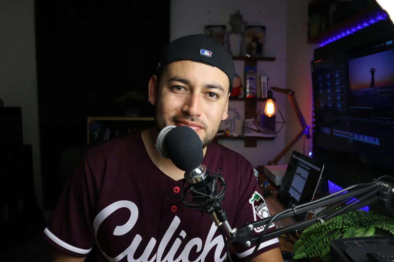 Man wearing a dark baseball cap and "Culich" jersey speaks into a microphone in a cozy, dimly lit room with a computer, plants, and soft lighting.