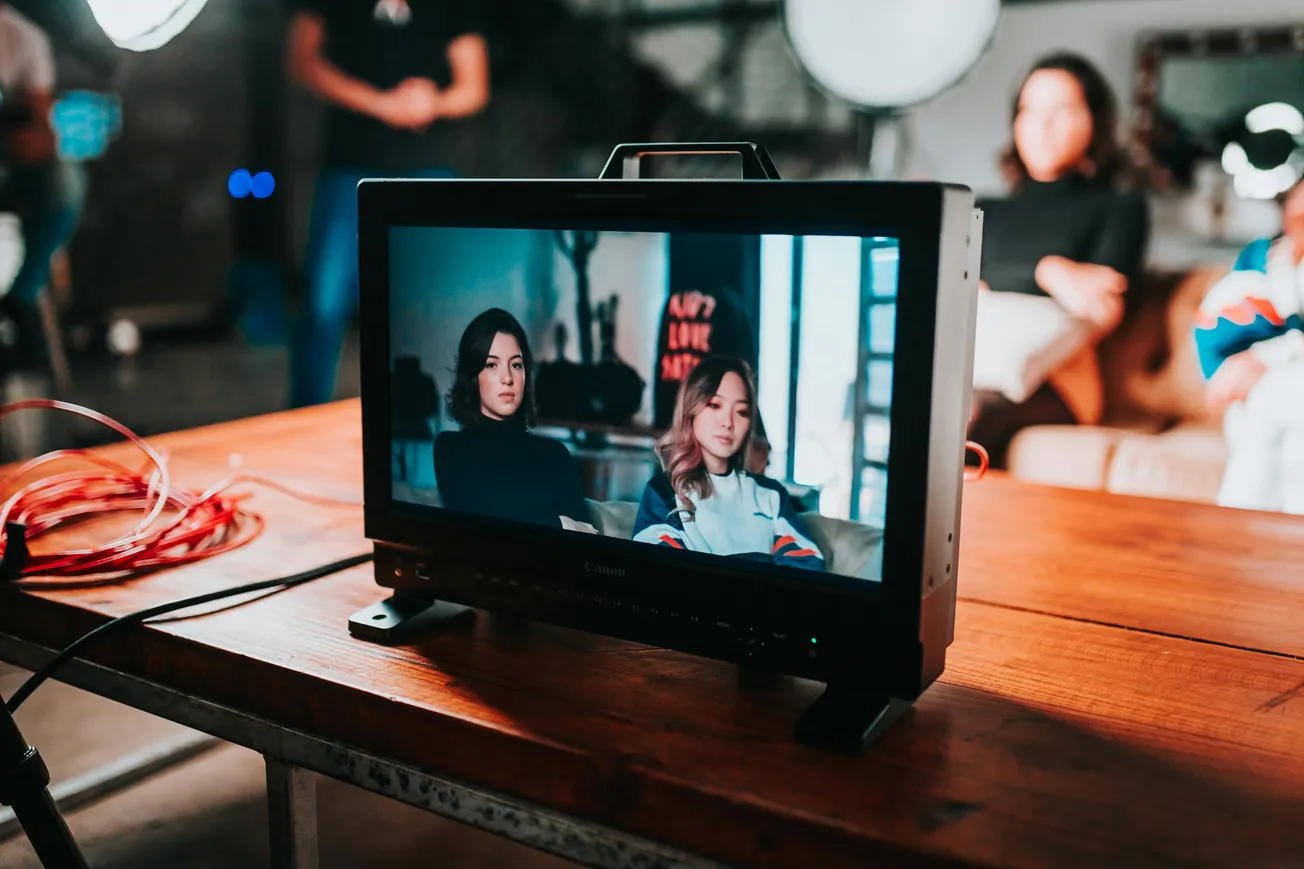 A monitor on a wooden table displays two women sitting, focused and serious. In the background, figures and studio lights are blurred, creating a professional film set ambiance.