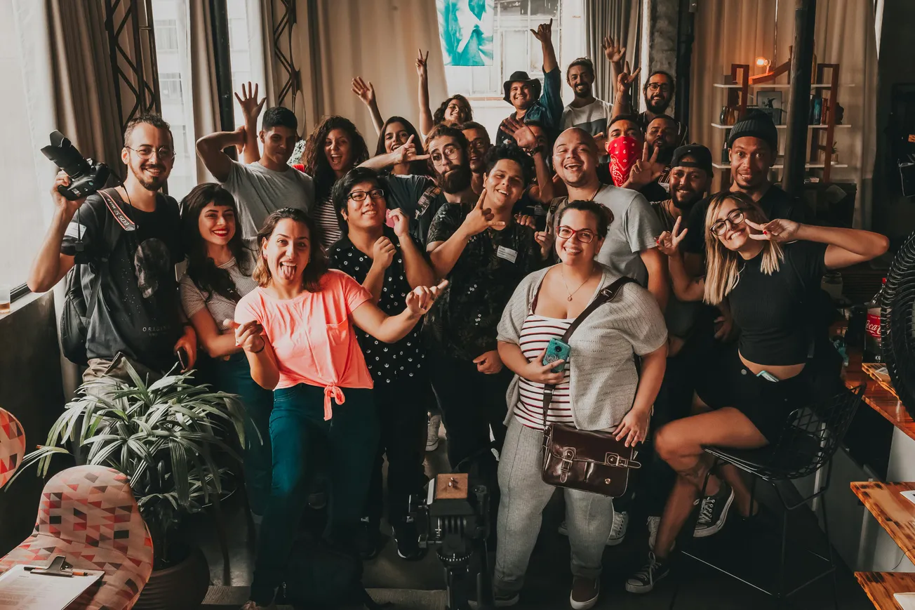A joyful group of diverse people posing indoors, smiling and gesturing peace signs. The setting is casual, with natural light from large windows.