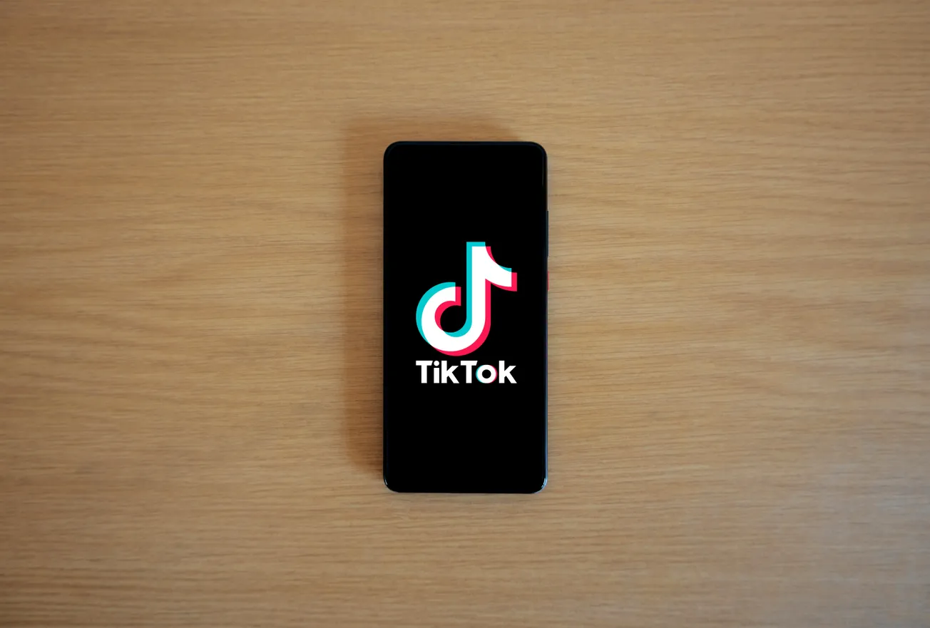 A smartphone displaying the TikTok logo, set on a light wooden surface. The black screen highlights the colorful logo, conveying a modern and digital theme.