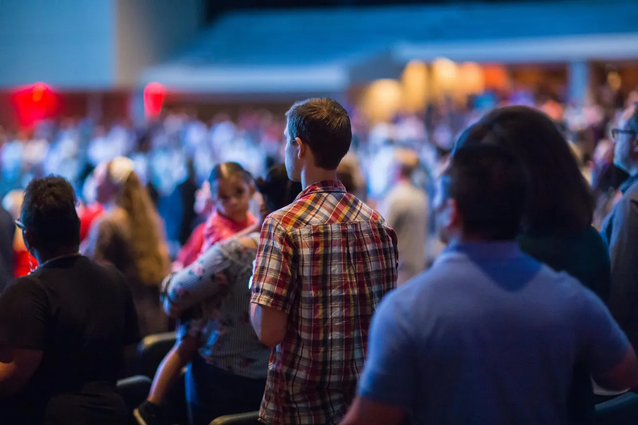A crowd of people standing indoors, focused on an event. The scene is lit with blue and warm tones, creating an attentive and lively atmosphere.