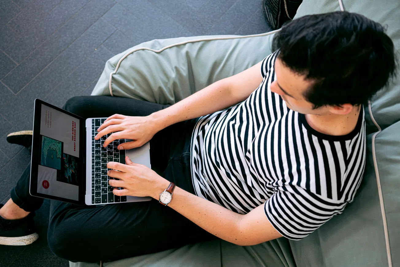 A person in a striped shirt is lounging on a beanbag while typing on a laptop. The relaxed setting and casual attire suggest a comfortable, work-from-home environment.