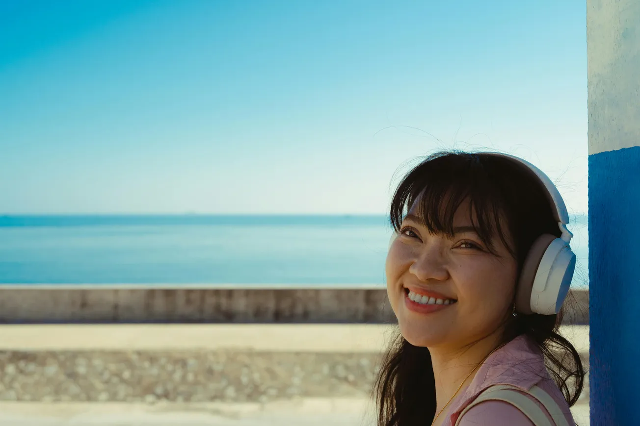 Woman smiling by the sea, wearing headphones. She leans against a blue wall, with the ocean and clear sky creating a peaceful, joyful scene.