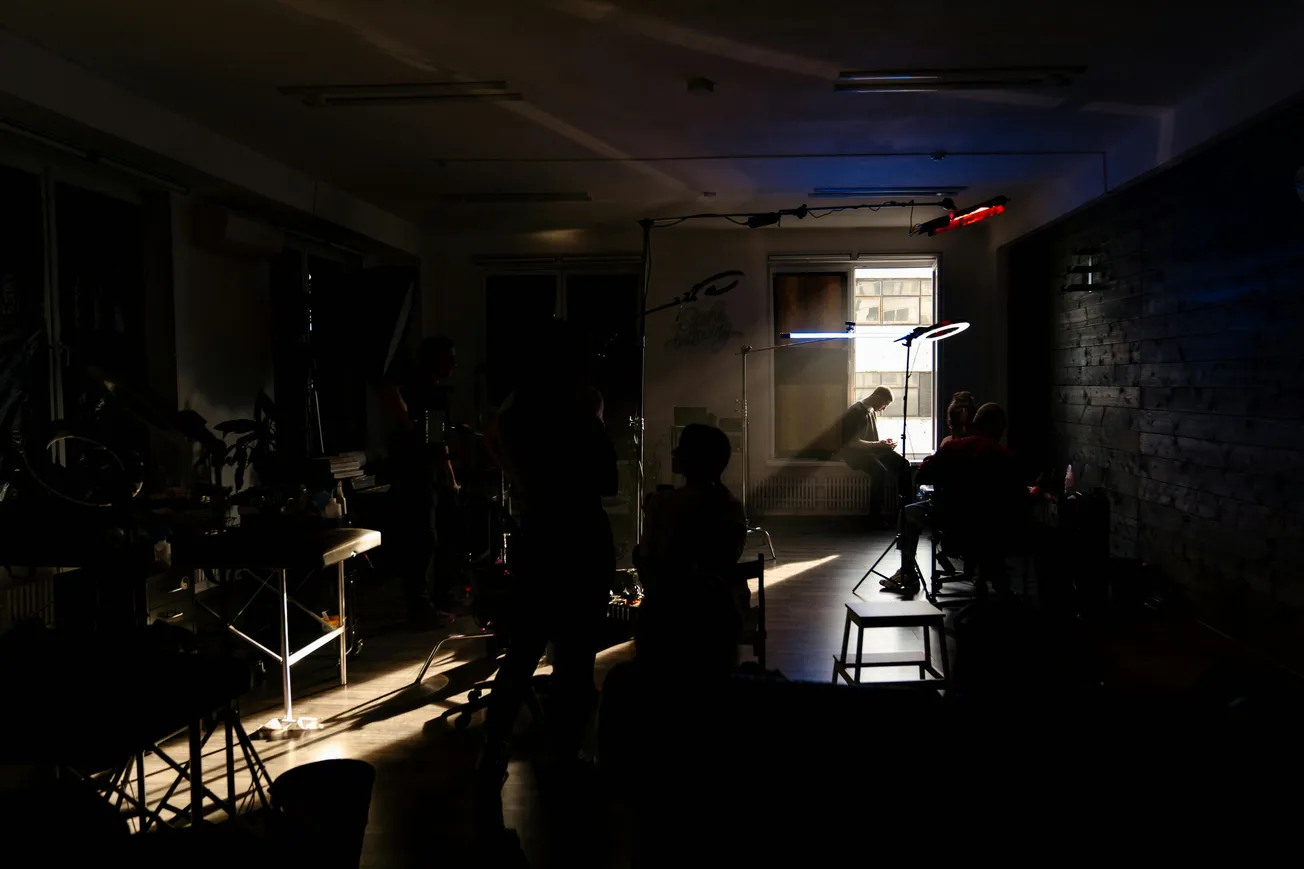 A dimly lit room with people silhouetted around filming equipment, focused on a person sitting at a window with natural light highlighting them. The scene feels intimate and cinematic.