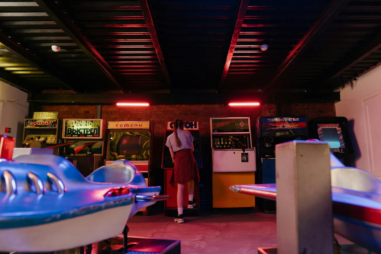 A person in a patterned red skirt and sneakers plays a vintage arcade game in a dimly lit room with neon lights and retro game machines. The atmosphere is nostalgic.