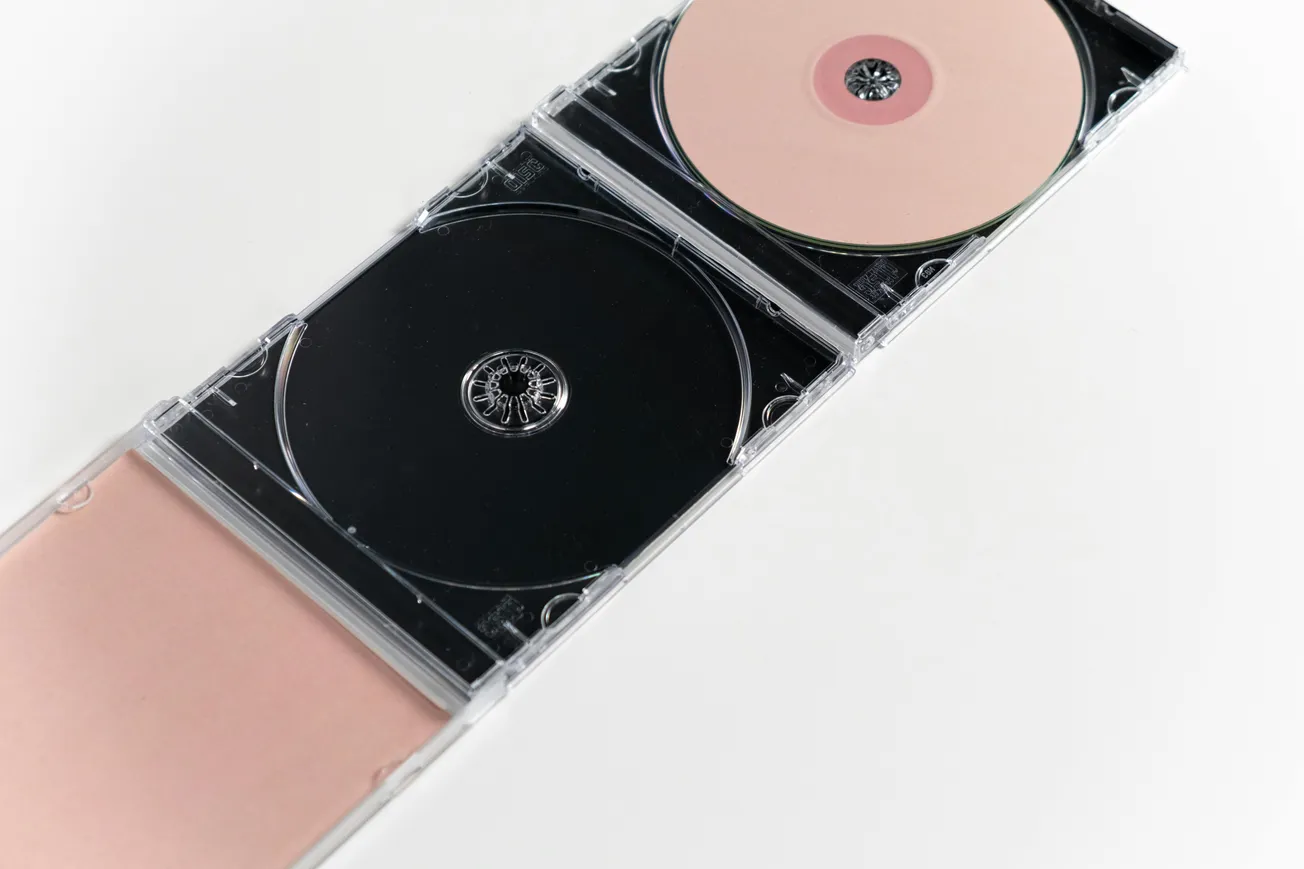 Open CD case on a white background, containing two disc slots. One disc is light pink with a darker pink center, and the other disc slot is empty..