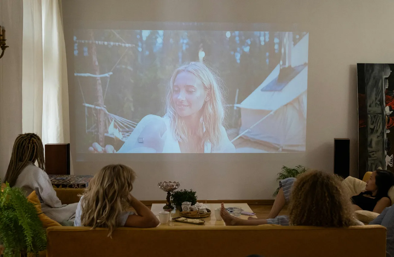 Four people sit on a sofa, watching a movie projected on a wall. The scene is cozy, with plants and warm lighting, creating a relaxed atmosphere.