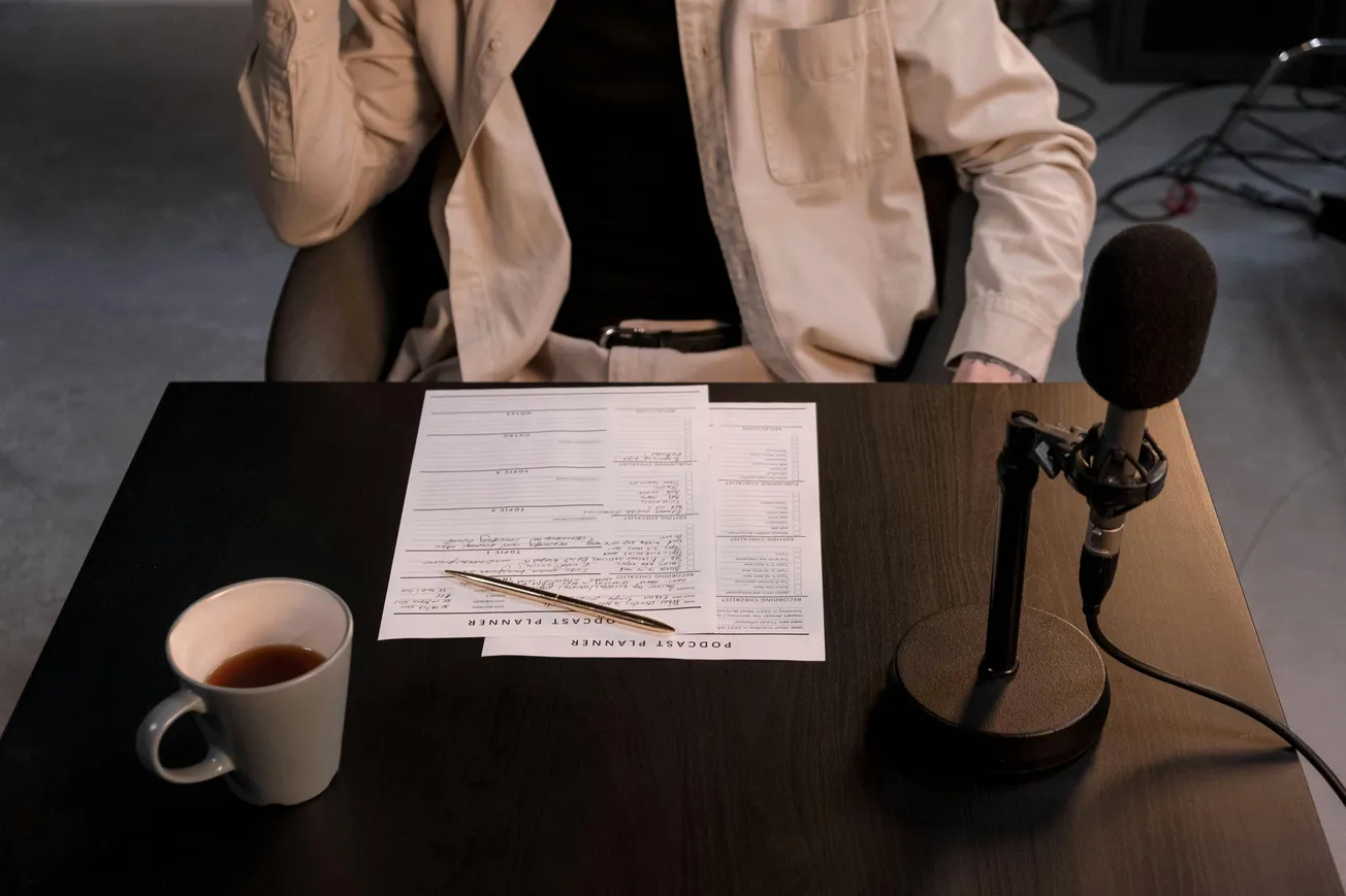 A person in a beige jacket sits at a table with a microphone, podcast planner, pencil, and coffee cup. The scene conveys a podcast recording setup.