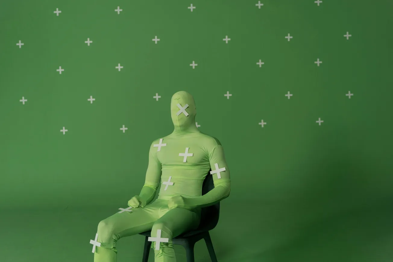 A person in a green morph suit sits against a green screen backdrop, marked with tracking crosses for visual effects, creating an artificial and neutral tone.