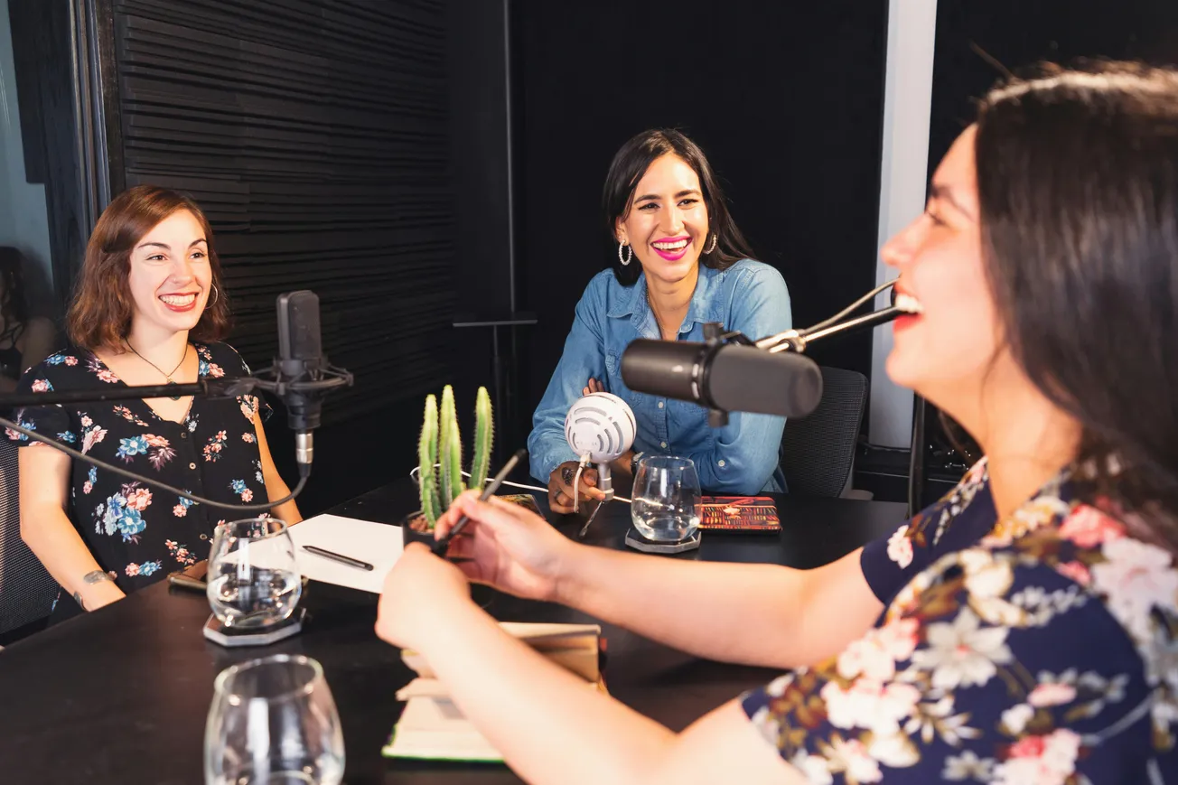 Three women laugh and talk around a podcast table with microphones and glasses of water. The setting is casual and friendly, conveying joy.