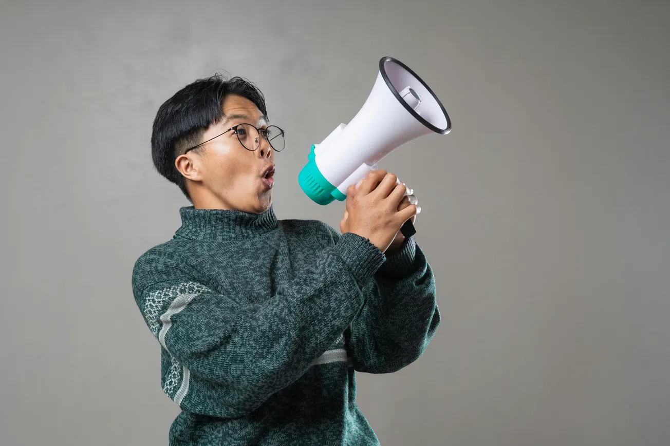 A person in glasses and a green sweater looks surprised while holding a white megaphone with teal accents against a gray background.