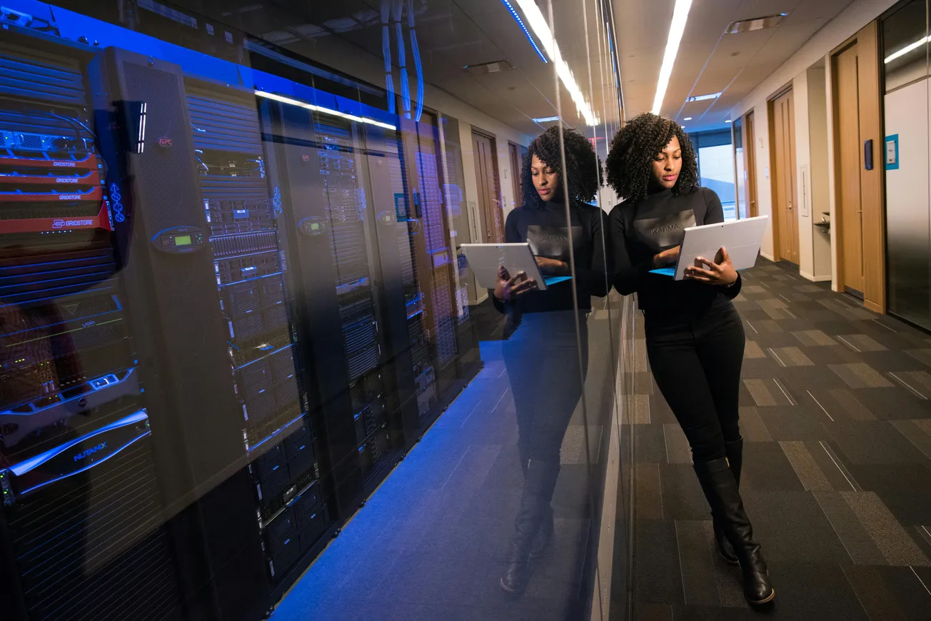 A woman leans on a glass wall in a server room, focused on her laptop. Blue light from server racks reflects, creating a tech-centric, modern vibe.