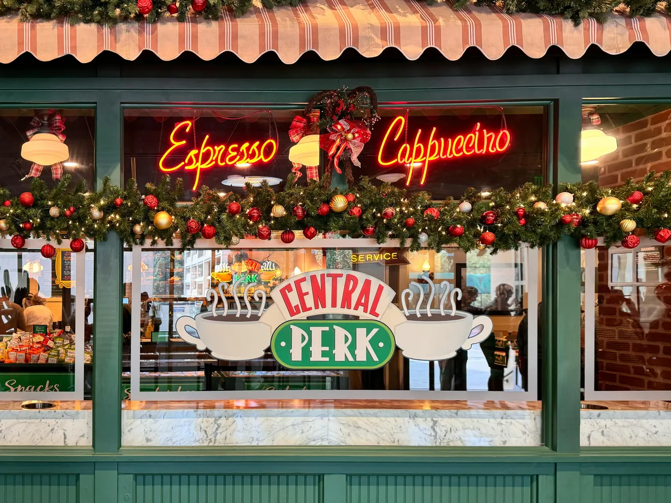Front of a coffee shop decorated for the holidays with garlands and ornaments. "Central Perk" logo on window; neon "Espresso" and "Cappuccino" signs. Cozy and festive mood.