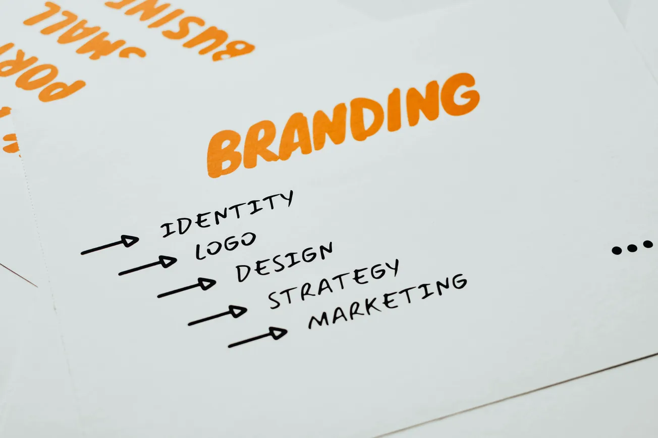 White paper showing the word "BRANDING" in bold orange text. Below, arrows point to words: Identity, Logo, Design, Strategy, Marketing.