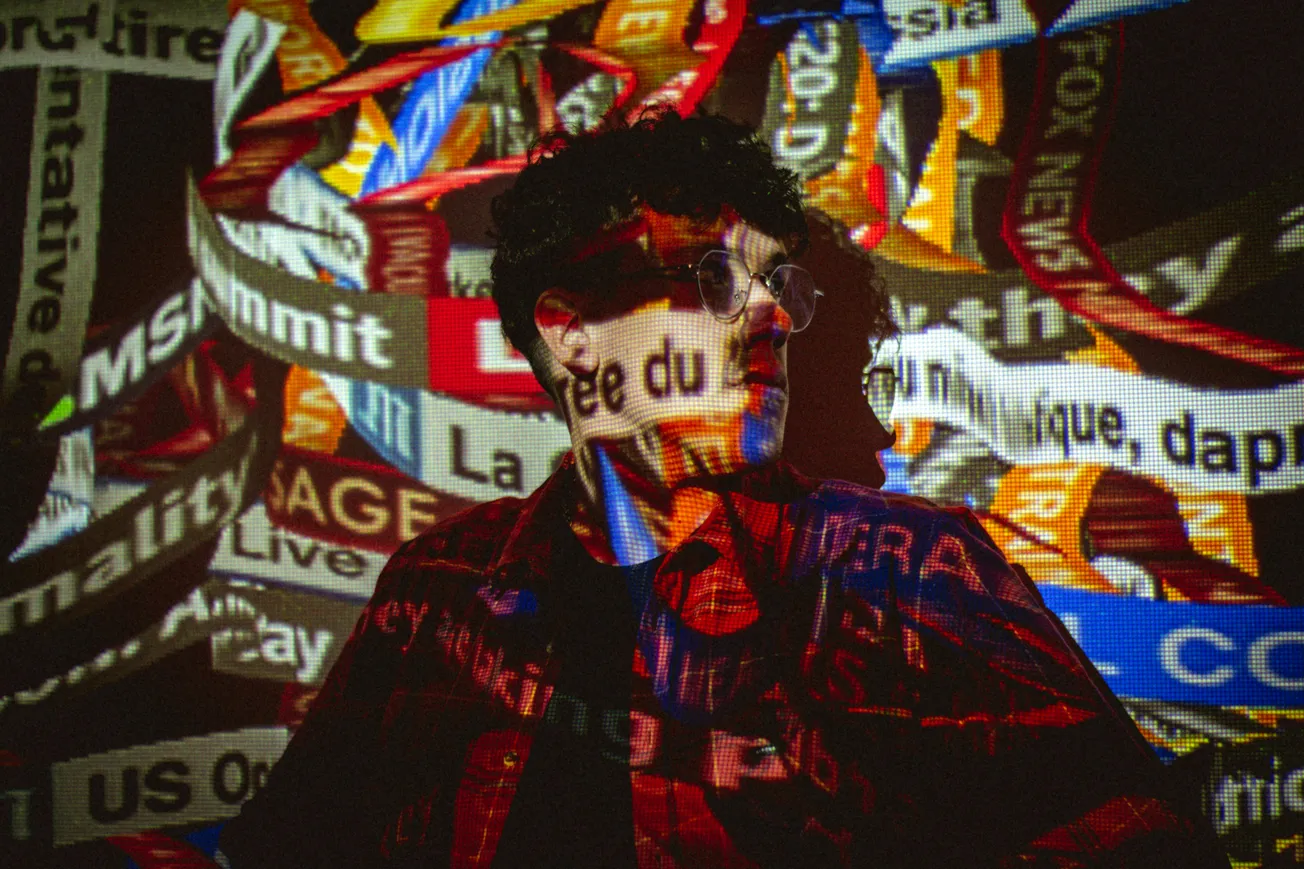 A person in glasses stands in front of a vibrant projection of colorful news headlines, creating a dynamic, layered effect on their face and clothing.