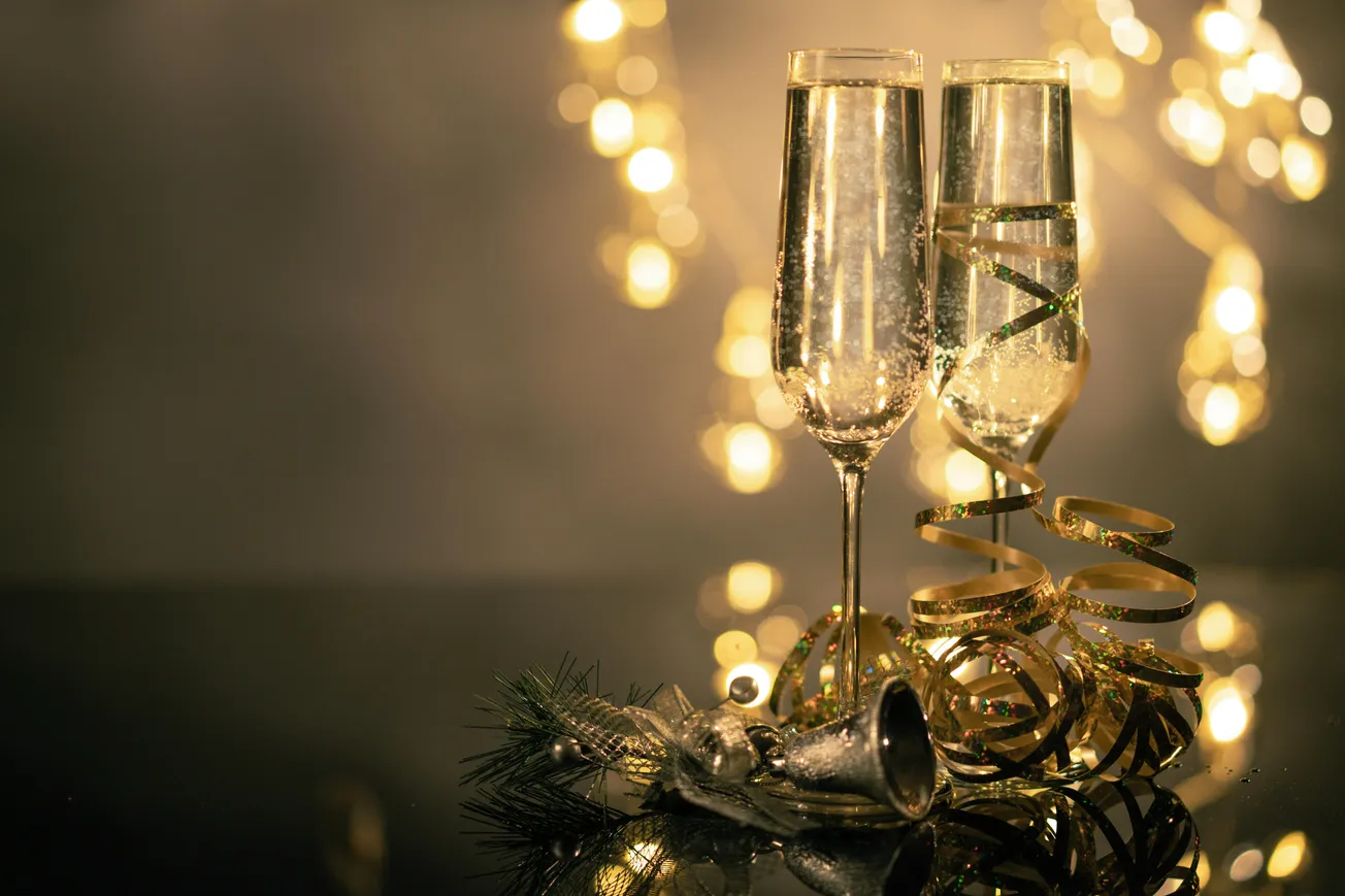Two champagne flutes with golden ribbon and festive lights create a celebratory mood. A pine cone and silver bell add a holiday touch.