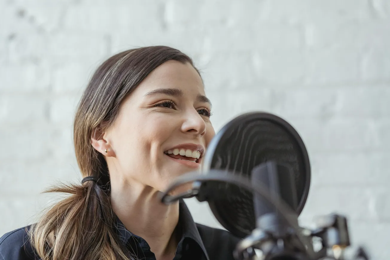 A woman with long brown hair smiles while speaking into a microphone with a pop filter. She appears joyful and engaged, set against a light brick wall.